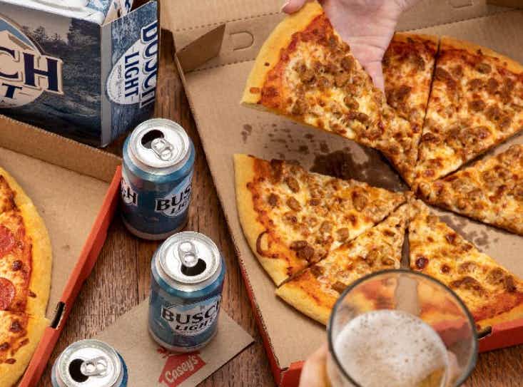 Sausage pizza from caseys next to a few beers with a person's hand grabbing a slice of pizza. 
