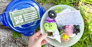 Someone picking up a Geocache Tupperware container with a bunch of items inside, including a Travel Bug with an Airtag attached