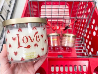 A Love candle held out by hand in front of a store cart with other candles lying in it.