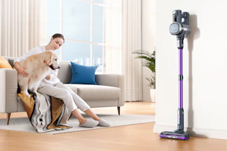 A woman sits on her beige couch next to a medium sized dog. The Lubluelu cordless vacuum cleaner stands on its own in the corner.