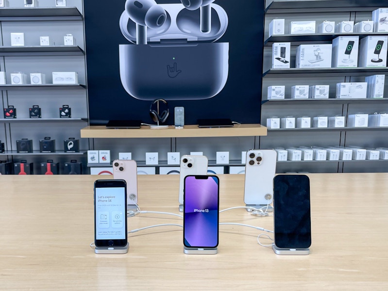 A variety of iPhones standing on a table with other apple products in the background.