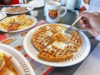 A person's hand using a fork to pick up a piece of a waffle from a plate on a Waffle House table next to other plated breakfast items.