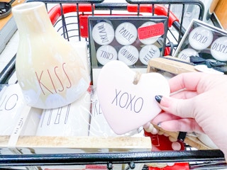 A person's hand holding up a Rae Dunn product in front of a shopping cart full of other Rae Dunn products in TJ Maxx.