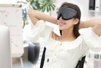 A woman in a white blouse sits at a desk wearing the heated eye mask and smiling.