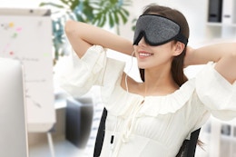 A woman in a white blouse sits at a desk wearing the heated eye mask and smiling.
