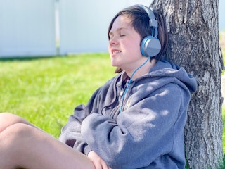a teen shiting in the grass leaning on a tree listening to music with headphones on