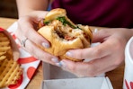 A person holding a Chick-fil-A sandwich with a bite taken out of it next to a container of waffle fries on a table in Chick-fil-A.