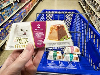person holding a container of fancy feast cat food over a blue cart with more cat food in it
