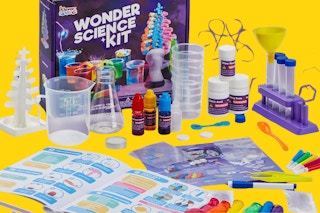 A kids' science kit has its contents spread on a yellow background. They include test tubes, beakers, food color, balloons, and more.