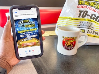 A person's hand holding a cell phone displaying a welcome email from Waffle House Regular Club in front of a Waffle House mug and to-go b...