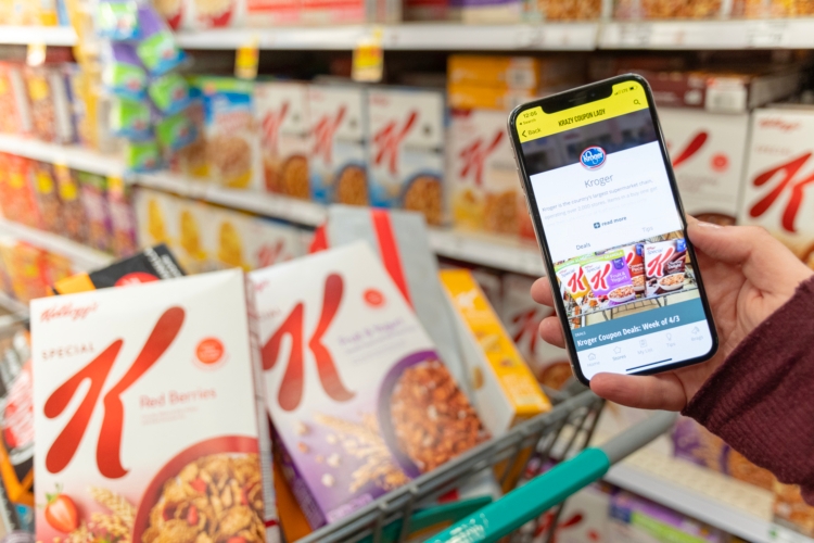 A person holding a smartphone standing next to a grocery cart with boxes of Special K cereal in it.