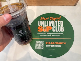 A person's hand holding a Panera drink cup next to a sign for their Unlimited Sip Club.