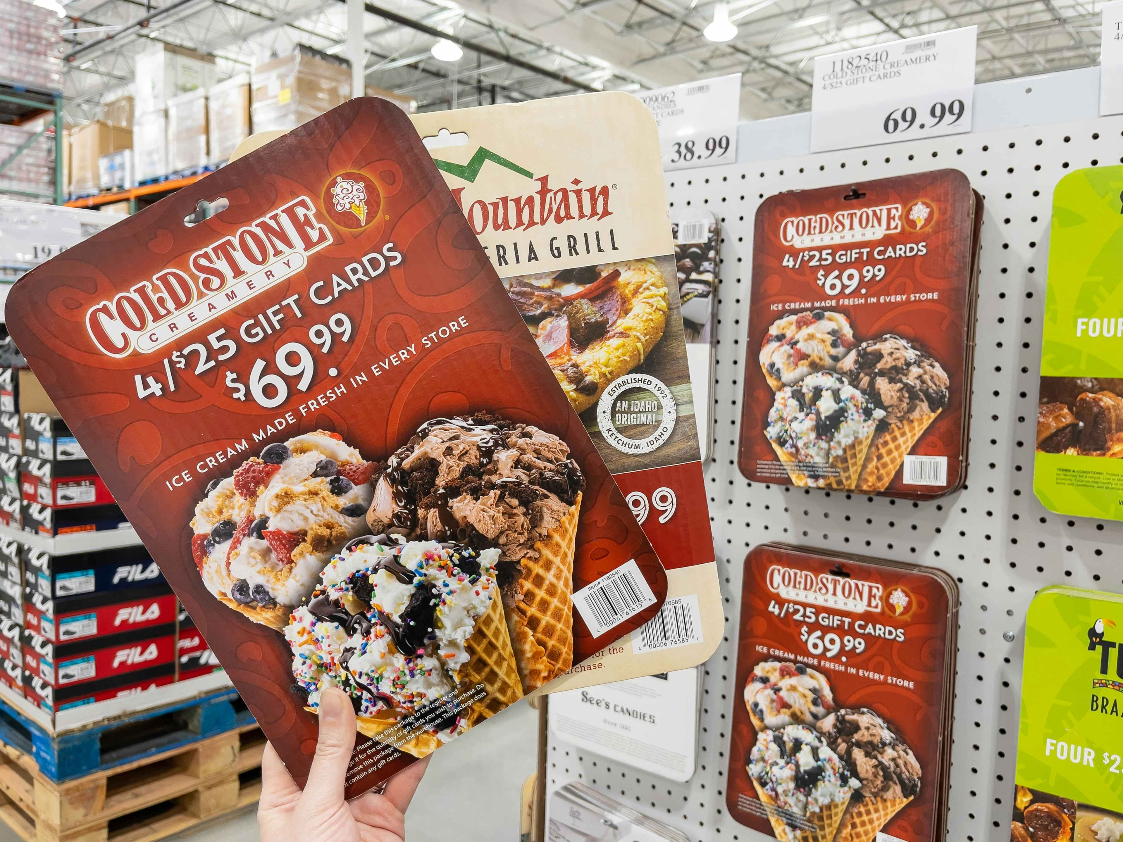 A person holding up two cardboard display cutouts for gift cards sold at Costco. One of the cards is for 4, $25 Cold Stone Creamery gift ...