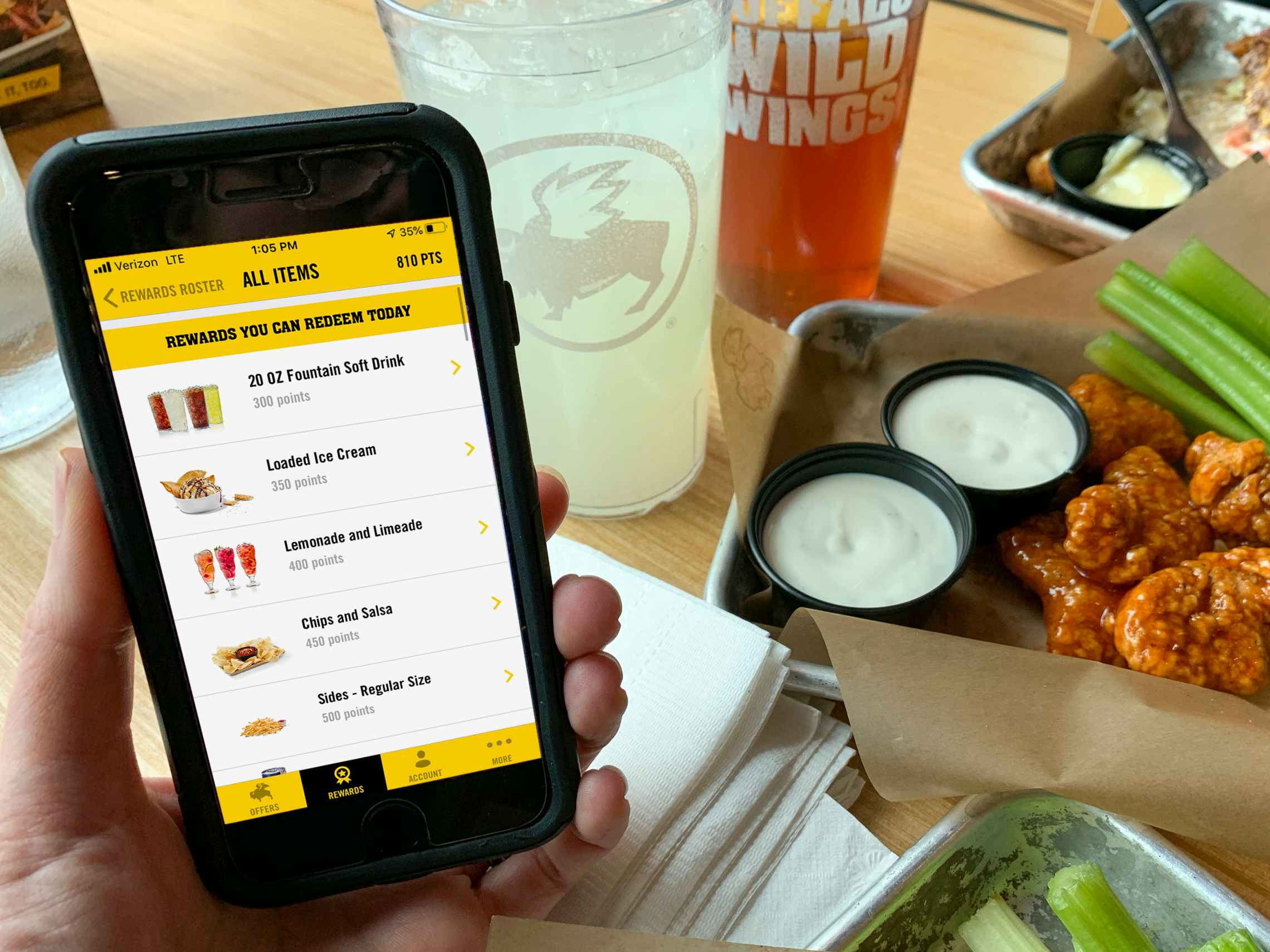 A person holding their iPhone with the Buffalo Wild Wings mobile app open to the Rewards Roster page; the list shows the user's redeemabl...