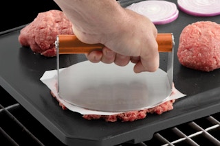 Cuisinart 6" Stainless Steel Smashed Burger Press being used by a person on the grill.