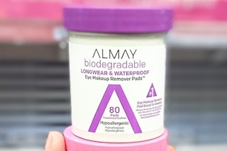 hand holding two Almay eye makeup remover jars in front of an Almay display in a store