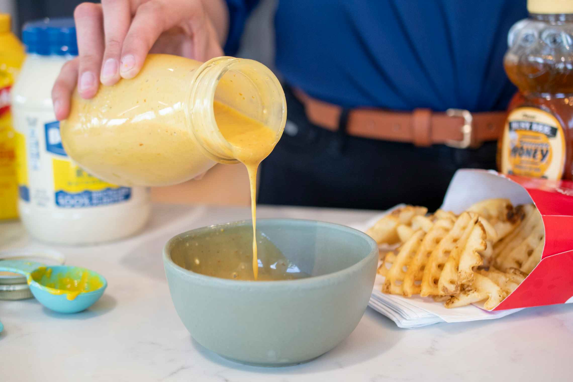 Homemade chick-fil-a sauce being poured from a glass bottle into a bowl on a table next to Chick-fil-A waffle fries and sauce ingredients...