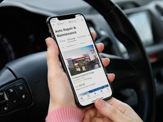 A person sitting in their car, holding a cell phone displaying the Auto Repair & Maintenance page with oil change offers on the Groupon app.