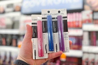hand holding three covergirl mascaras in different colors in front of a covergirl makeup display