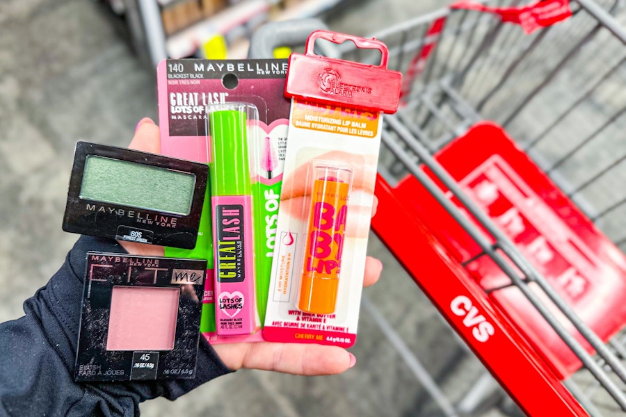 person holding maybelline baby lips, mascara, blush, and eyeshadow over a cvs cart