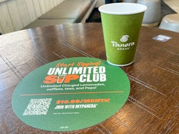 A panera bread cup next to an unlimited sip club sticker on a table.