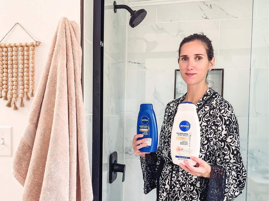 A woman wearing a bathrobe, standing in front of a walk in shower, holding two bottles of Nivea shower products.