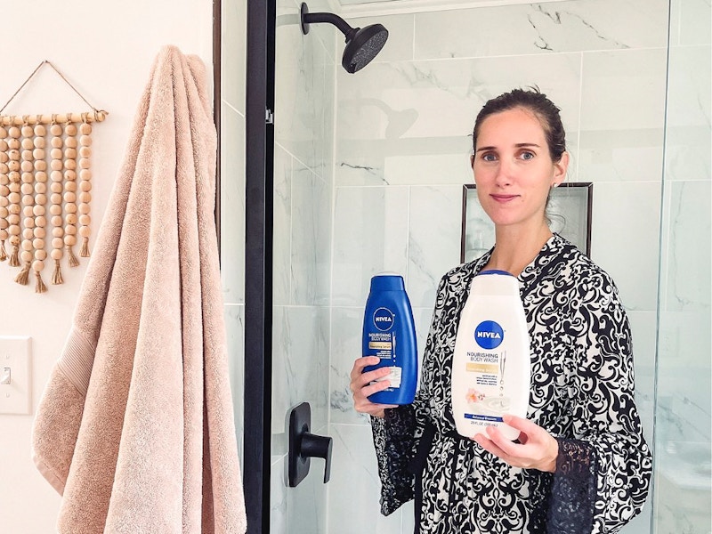 A woman wearing a bathrobe, standing in front of a walk in shower, holding two bottles of Nivea shower products.