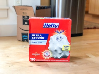 A box of Hefty trash bags sitting on the floor of a kitchen with an Amazon box behind it.