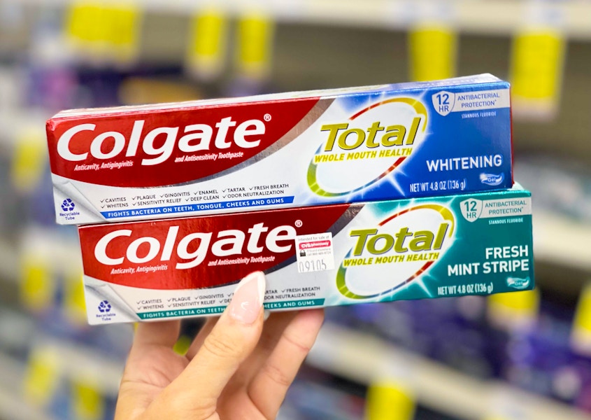 A person's hand holding up two stacked boxes of Colgate Total brand toothpaste in front of a shelf at CVS.