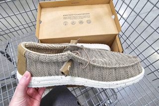 person holding a brown hey dude shoe over a cart with the shoe box in it