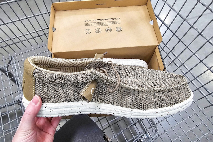 person holding a brown hey dude shoe over a cart with the shoe box in it
