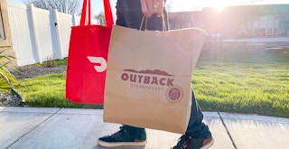 a person holding a DoorDash delivery bag and an Outback Steakhouse takeout bag walking down a sidewalk
