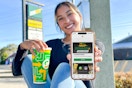 a woman holding a cellphone with the subway app and a subway drink sitting outside a subway 