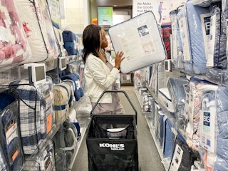 A woman standing behind a shopping basket pulling a bedding set off the shelf at Kohl's