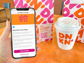 A person's hand holding an iPhone displaying the welcome to DDPerks page on the Dunkin app next to a small coffee and a Dunkin bag inside...