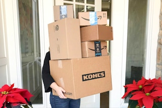 A person standing in their front doorway holding a very tall stack of packages