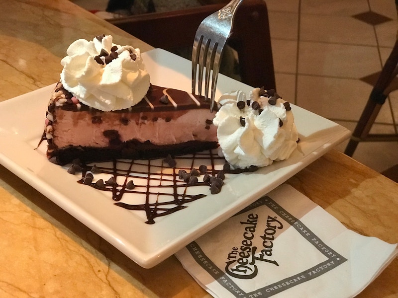 A plate with chocolate cheesecake with whipped cream and a cheesecake factory napkin next to it on a table.