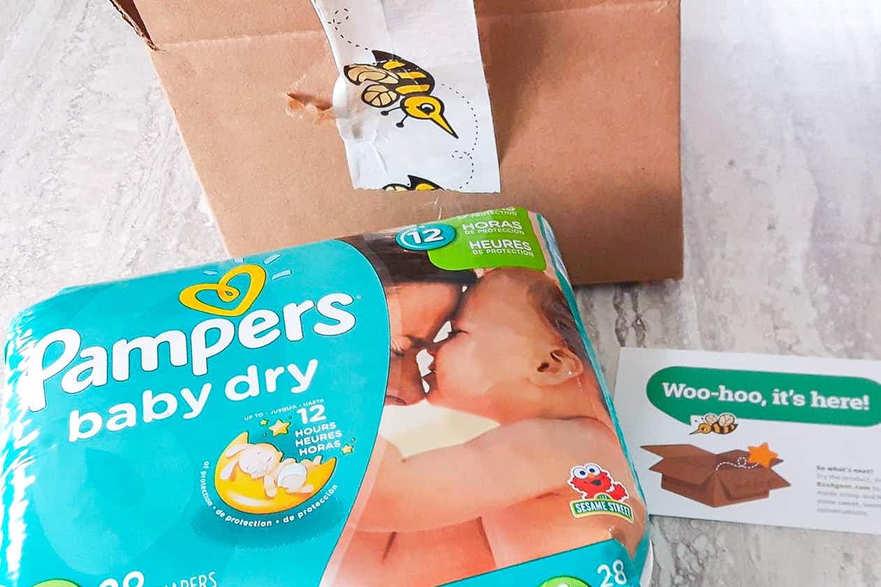 A package of Pampers diapers on a table next to a shipping box from BzzAgent and a card that reads, "woo-hoo, it's here!"