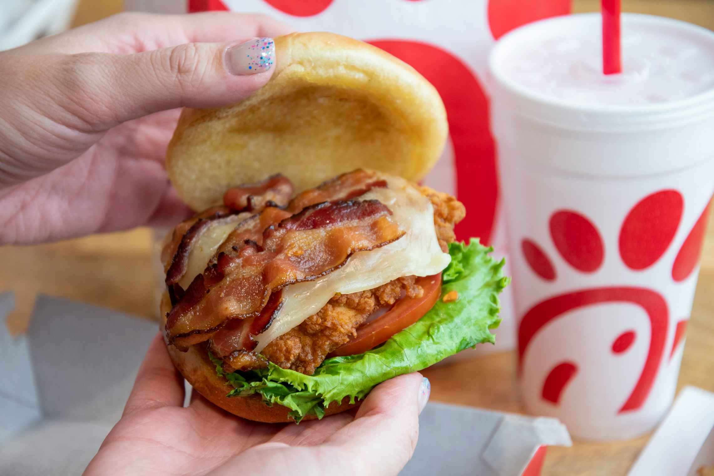 A person's hands lifting the top bun off of a Chick-fil-A chicken sandwich with bacon to show the ingredients. On the table in the backgr...