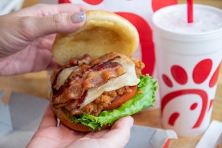 A person's hands lifting the top bun off of a Chick-fil-A chicken sandwich with bacon to show the ingredients. On the table in the backgr...