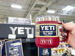 A person's hand holding a Yeti Rambler mug next to a Yeti display inside a store.