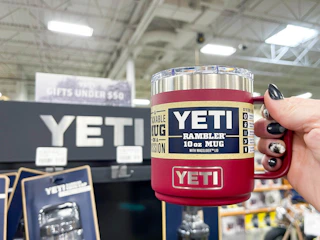 A person's hand holding a Yeti Rambler mug next to a Yeti display inside a store.