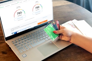 A person using a laptop displaying the Credit Karma website and holding a handful of credit cards.