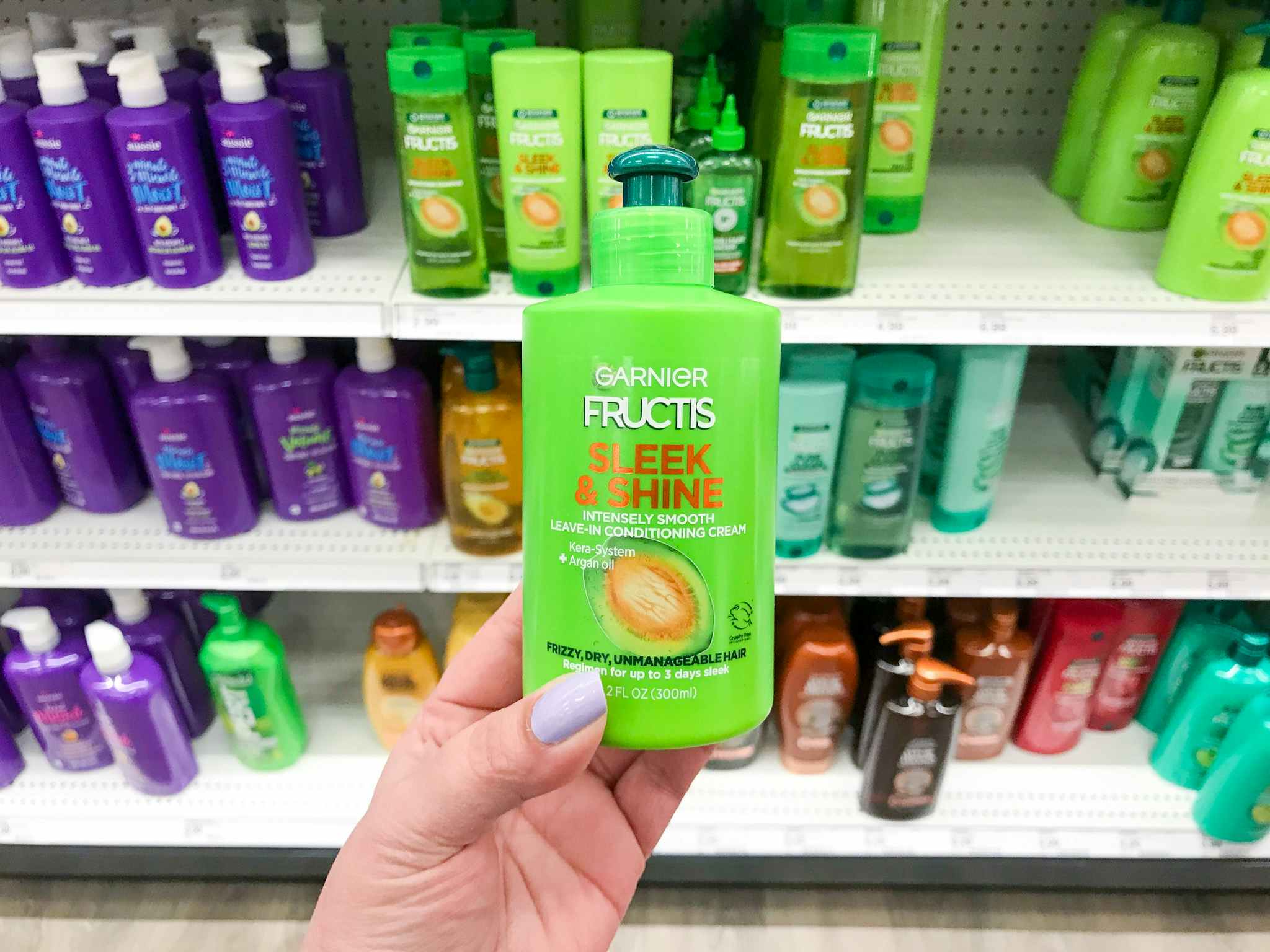 A person's hand holding up a bottle of Garnier Fructis Sleek & Shine Leave-in Conditioner in front of a shelf of haircare products at ...