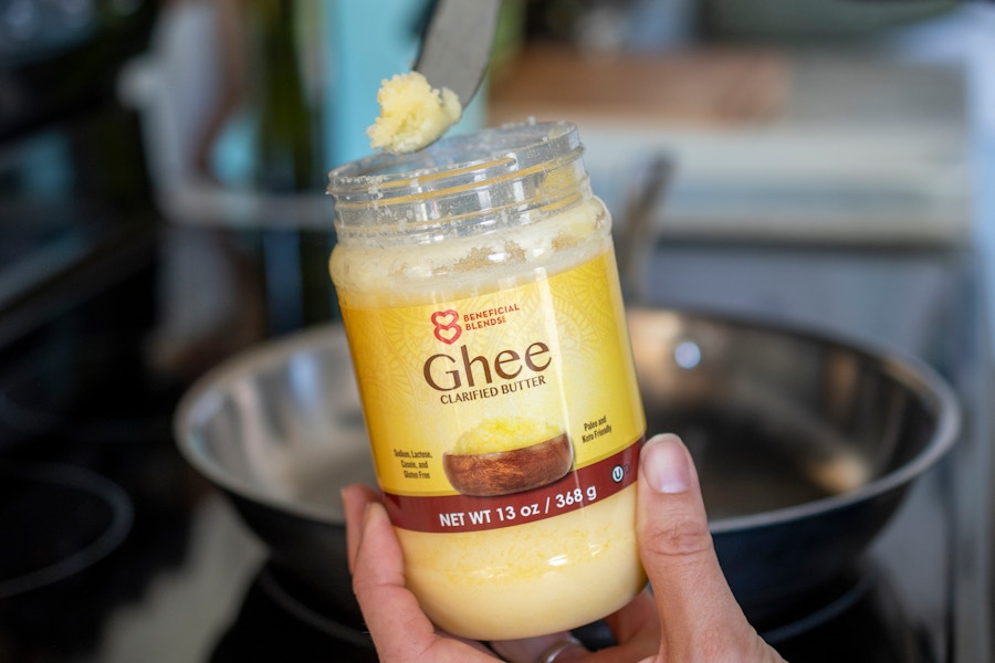 A person scooping Ghee out of its container next to a stove with a frying pan in the background.