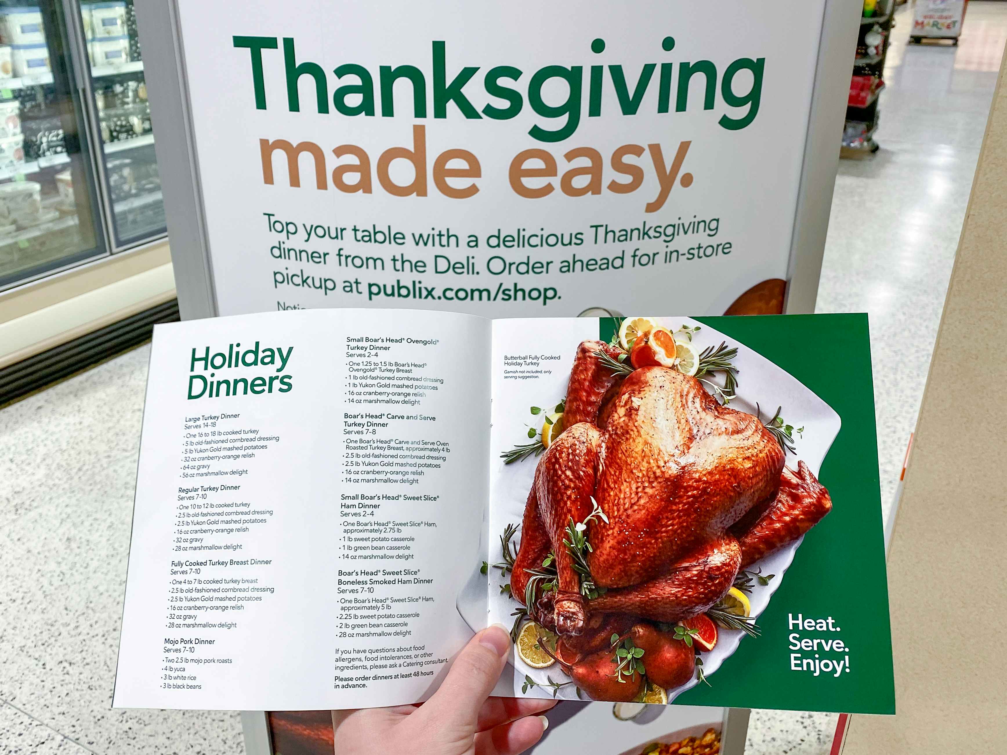 A person holding a booklet listing the Publix holiday dinner choices in front of a sign inside Publix that says, "Thanksgiving made easy....