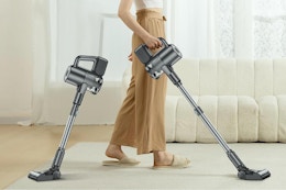 the bottom half of a person using a cordless stick vacuum cleaner. Next to them is the same vacuum but shorter