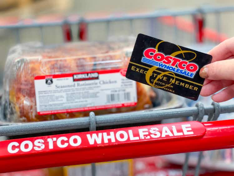 Hand holding Costco Executive Membership card in front of rotisserie chicken in shopping cart