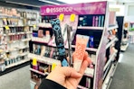 person holding essence mascara, eyeliner, and lip gloss in front of a makeup display