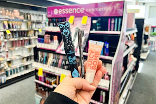 person holding essence mascara, eyeliner, and lip gloss in front of a makeup display